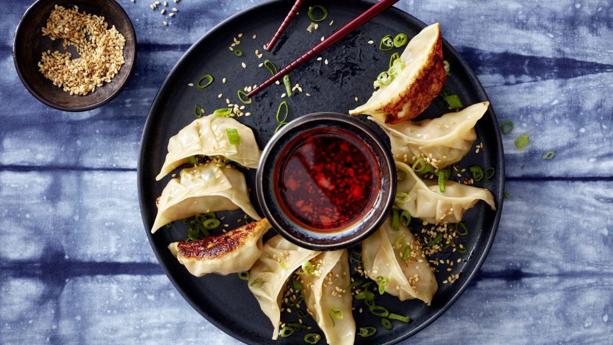 Ein schwarzer Teller mit den Garnelen-Dumplings auf einem jeansblauem Tisch mit Sesam. Alles direkt von oben fotografiert.