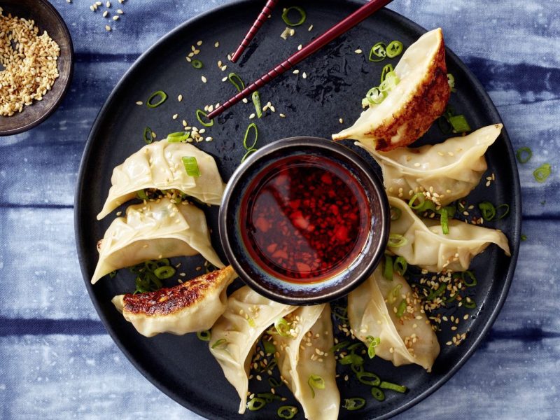 Ein schwarzer Teller mit den Garnelen-Dumplings auf einem jeansblauem Tisch mit Sesam. Alles direkt von oben fotografiert.