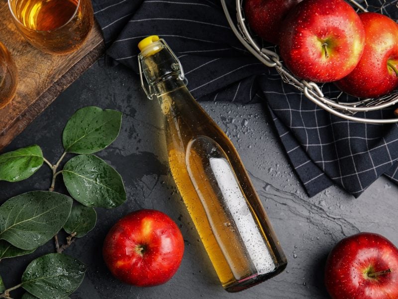 Draufsicht: Auf dunklem Untergrund liegt waagerecht eine Flasche mit einer Flüssigkeit. Daneben liegen frische Äpfel und einige Kräuter am Stängel.