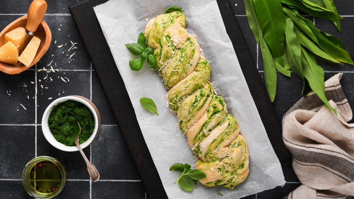 Draufsicht: Auf dunklem Untergrund sieht ein Backblech mit einem Kräuterbaguette. Drum herum liegen frische Zutaten, wie Bärlauch und Parmesanstücke.