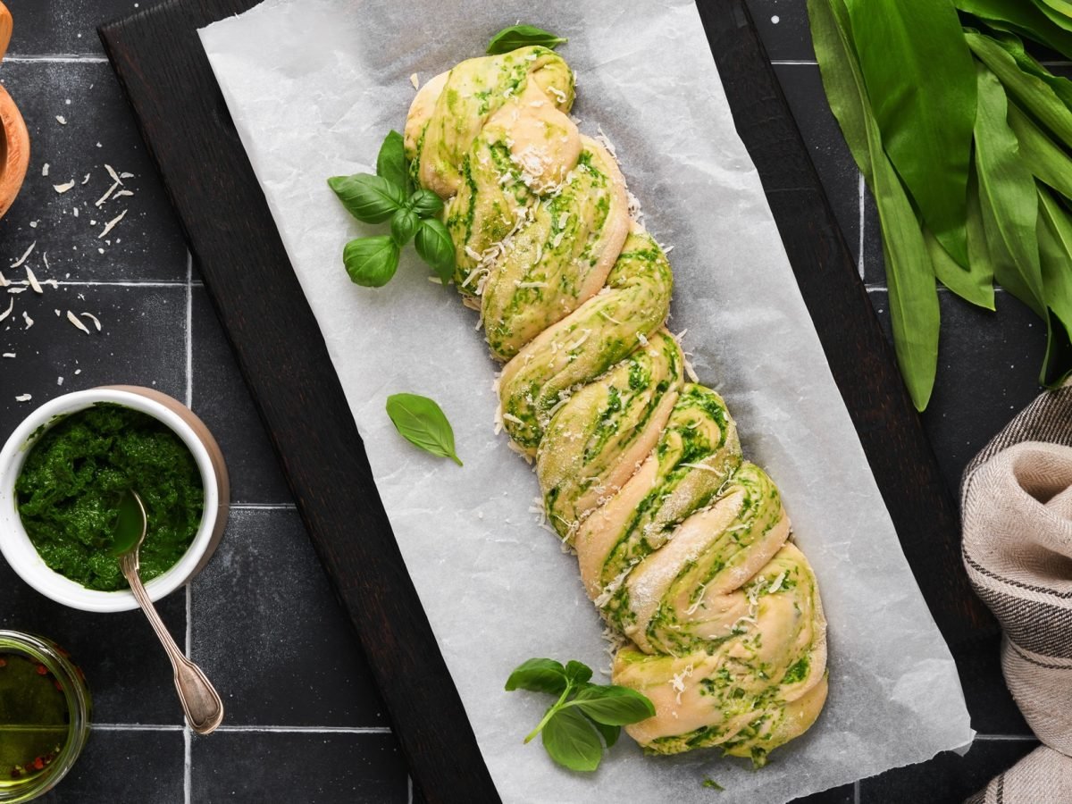 Draufsicht: Auf dunklem Untergrund sieht ein Backblech mit einem Kräuterbaguette. Drum herum liegen frische Zutaten, wie Bärlauch und Parmesanstücke.