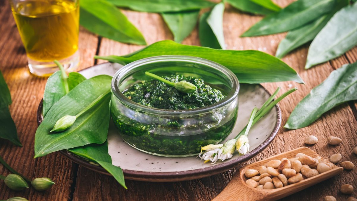 Draufsicht schräg: Ein Dip oder Aufstrich in einer Glasschüssel, der aus frischen Kräutern hergestellt wurde. drum herum liegen frische Bärlauchblätter und Pinienkerne.