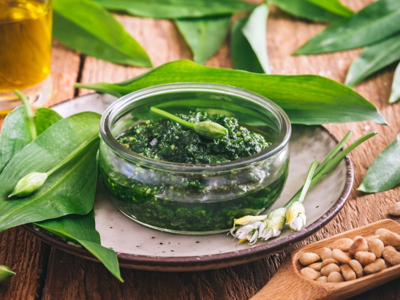 Draufsicht schräg: Ein Dip oder Aufstrich in einer Glasschüssel, der aus frischen Kräutern hergestellt wurde. drum herum liegen frische Bärlauchblätter und Pinienkerne.