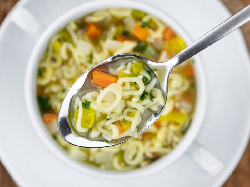 Draufsicht: Ein runder, heller Teller auf einem Holzboden. In dem Teller ist eine Buchstabensuppe mit Gemüse und Kräutern.
