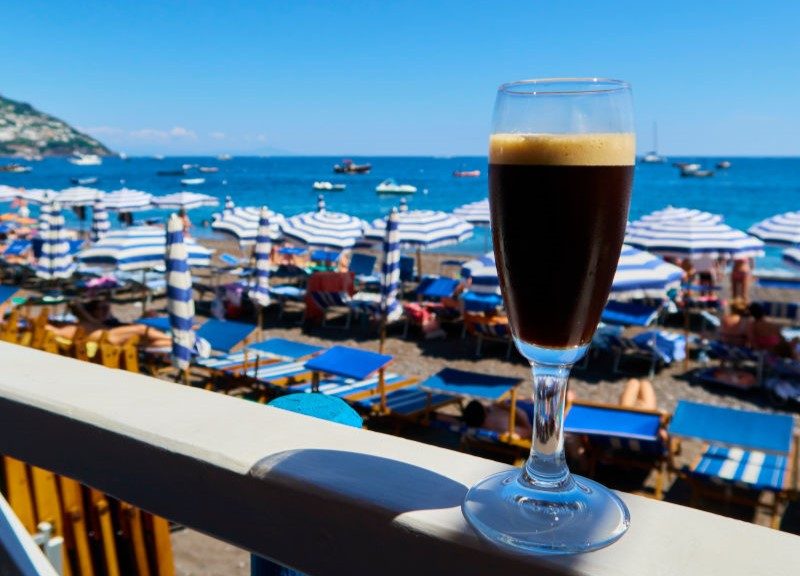 Frontal: Ein italienischer Strand mit vielen Sommerschirmen. Frontal steht ein Sektglas mit einer dunklen Flüssigkeit auf einem Geländer. In dem Glas ist ein Kaffee.