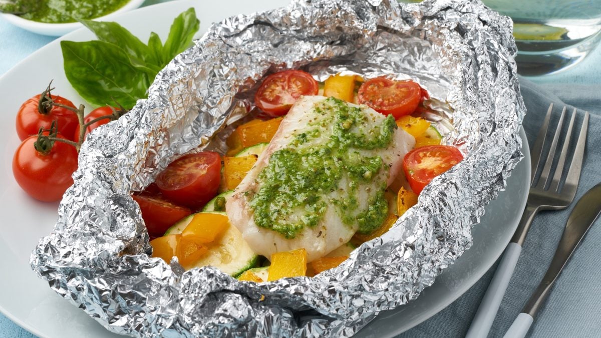 Draufsicht: Auf hellem Untergrund liegt eine geöffnete Alufolie. In der sind ein Fischfilet und frisches Gemüse. neben den Päckchen liegen frische Tomaten und ein paar Blätter Basilikum.