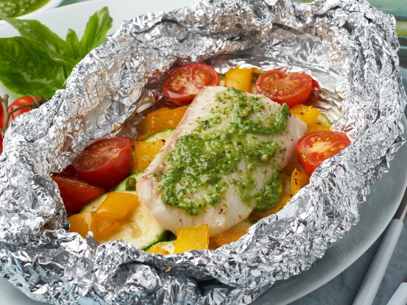 Draufsicht: Auf hellem Untergrund liegt eine geöffnete Alufolie. In der sind ein Fischfilet und frisches Gemüse. neben den Päckchen liegen frische Tomaten und ein paar Blätter Basilikum.