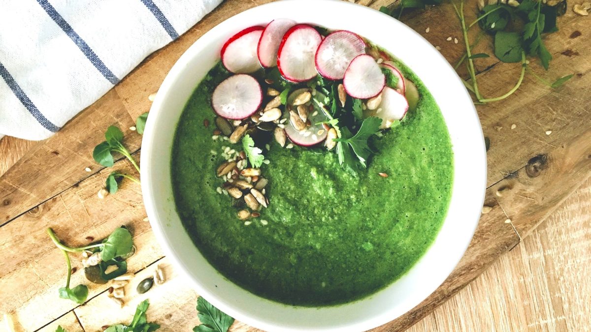 Draufsicht: Eine runde helle Schüssel mit einen Gemüsesuppe. Die ist mit Radieschen sowie Nüssen und Samen garniert.