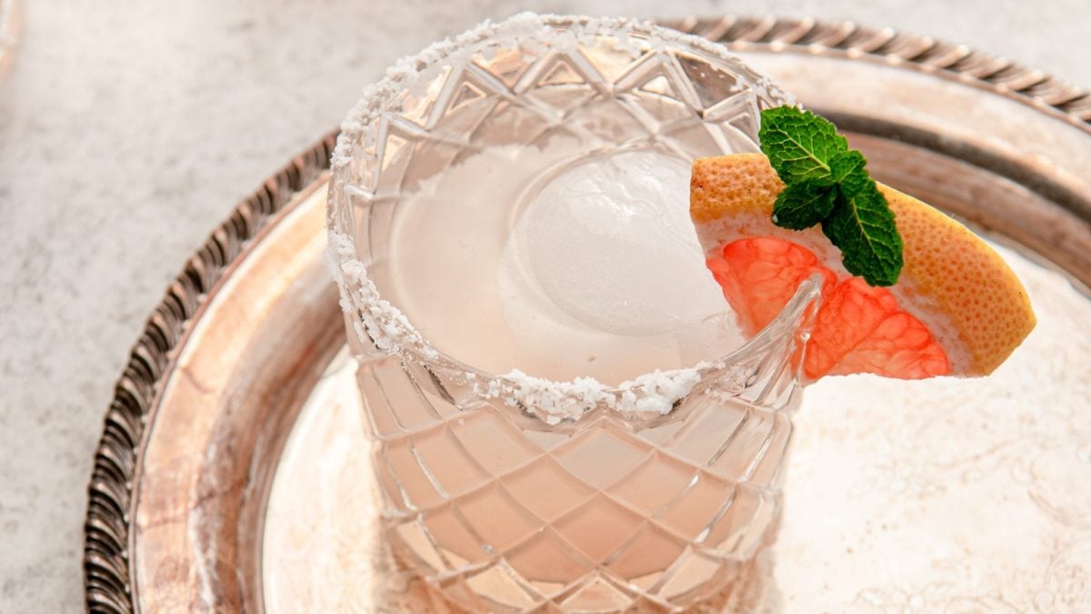 Draufsicht, schräg: Auf einem silbernen Tablett steht ein mit einer Flüssigkeit gefülltem Glas. Der Glasrand hat eine Salzkruste. Und in den Rand ist eine Scheibe eine Grapefruit als Dekoration eingeklemmt.