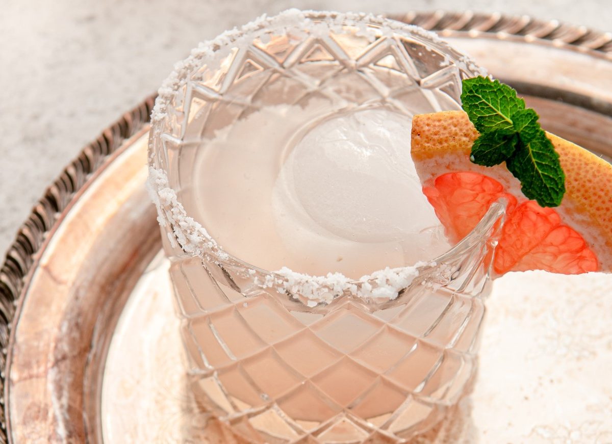 Draufsicht, schräg: Auf einem silbernen Tablett steht ein mit einer Flüssigkeit gefülltem Glas. Der Glasrand hat eine Salzkruste. Und in den Rand ist eine Scheibe eine Grapefruit als Dekoration eingeklemmt.