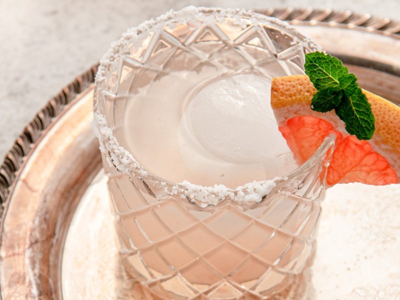 Draufsicht, schräg: Auf einem silbernen Tablett steht ein mit einer Flüssigkeit gefülltem Glas. Der Glasrand hat eine Salzkruste. Und in den Rand ist eine Scheibe eine Grapefruit als Dekoration eingeklemmt.