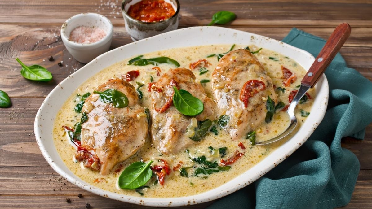 Draufsicht, schräg: Ein heller ovaler Teller aus Porzellan liegt auf einem Tisch aus Holz. Aus dem Teller sind drei Hähnchenfilets in einer Sauce mit Spinat und kleinen Tomaten. Hinter dem Teller stehen zwei kleine runde Gefäße aus Porzellan, in denen jeweils unterschiedliche Saucen sind.