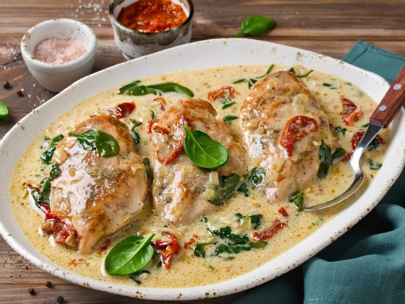 Draufsicht, schräg: Ein heller ovaler Teller aus Porzellan liegt auf einem Tisch aus Holz. Aus dem Teller sind drei Hähnchenfilets in einer Sauce mit Spinat und kleinen Tomaten. Hinter dem Teller stehen zwei kleine runde Gefäße aus Porzellan, in denen jeweils unterschiedliche Saucen sind.