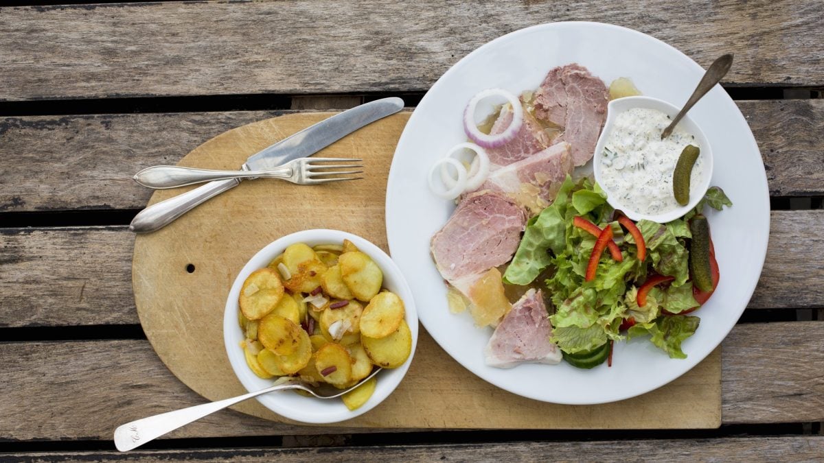 Draufsicht: Auf einem Untergrund aus Holz liegt ein rustikales Holzbrett auf dem eine weißer Teller mit einer Art Sülze, einer hellen Sauce und einige Salatblätter sowie eine runde helle Schüssel mit Bratkartoffeln liegen.