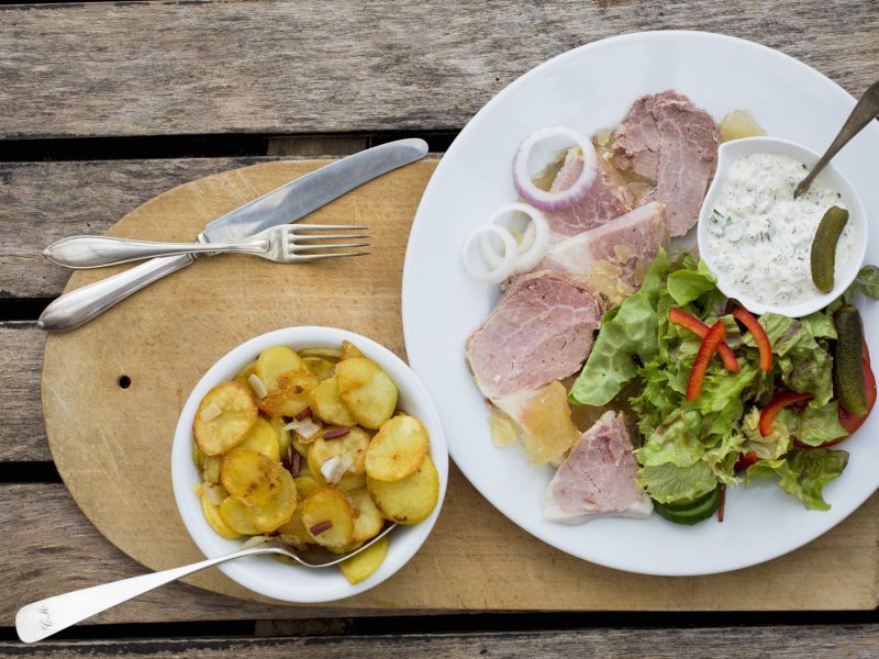 Draufsicht: Auf einem Untergrund aus Holz liegt ein rustikales Holzbrett auf dem eine weißer Teller mit einer Art Sülze, einer hellen Sauce und einige Salatblätter sowie eine runde helle Schüssel mit Bratkartoffeln liegen.