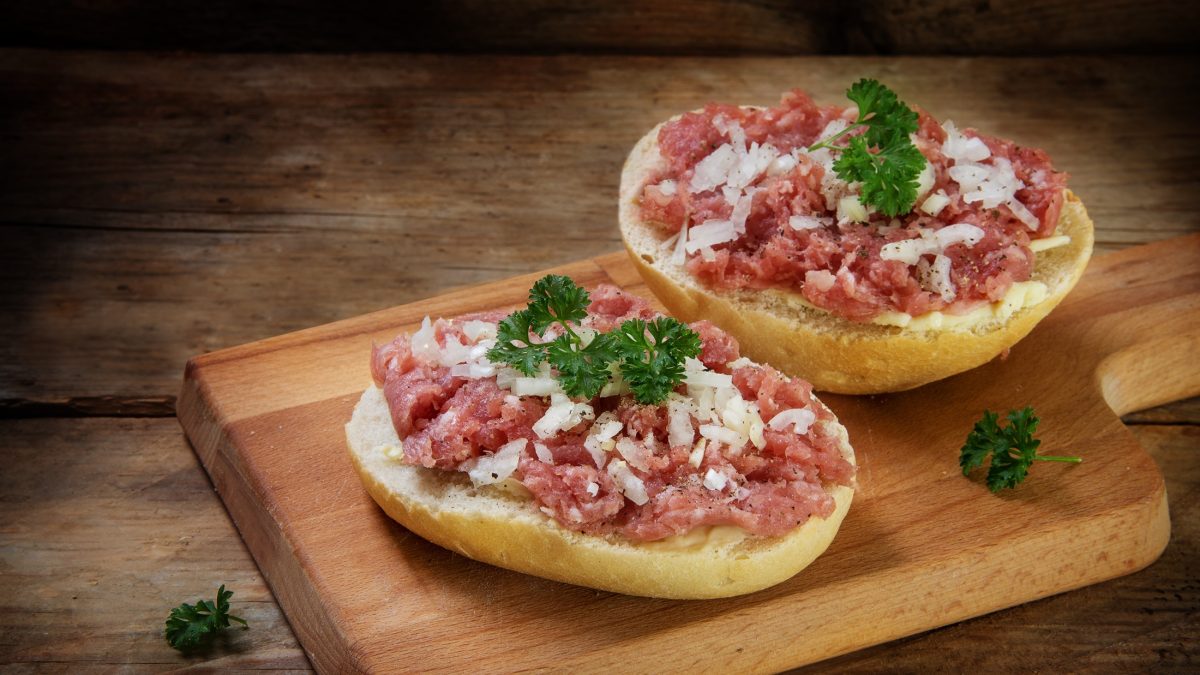 Draufsicht schräg: Auf einem Untergrund aus Holz liegen zwei Hälften Mettbrötchen mit Zwiebeln und Petersilie.