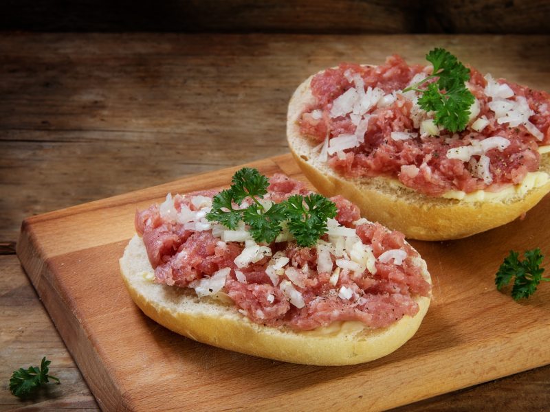 Draufsicht schräg: Auf einem Untergrund aus Holz liegen zwei Hälften Mettbrötchen mit Zwiebeln und Petersilie.