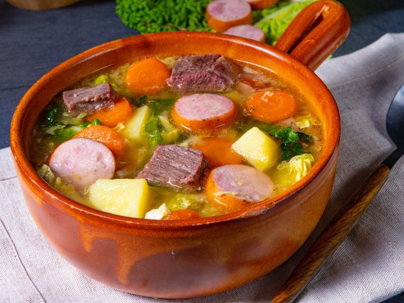 Draufsicht, schräge: Eine Suppenschale aus Ton auf einem hellen Tuch, dass auf einem dunklen Untergrund liegt. In der Schüssel ist ein Wirsing-Eintopf mit Möhren und Kartoffeln, Würstchen und Suppenfleisch.