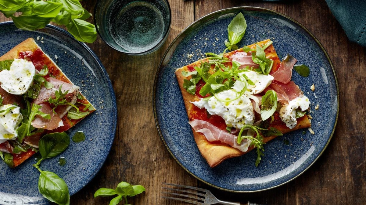 Pizza Crudo auf zwei grauen Tellern verteilt. Der Hintergrund ist hölzern, darauf Basilikum und ein Glas Wasser.