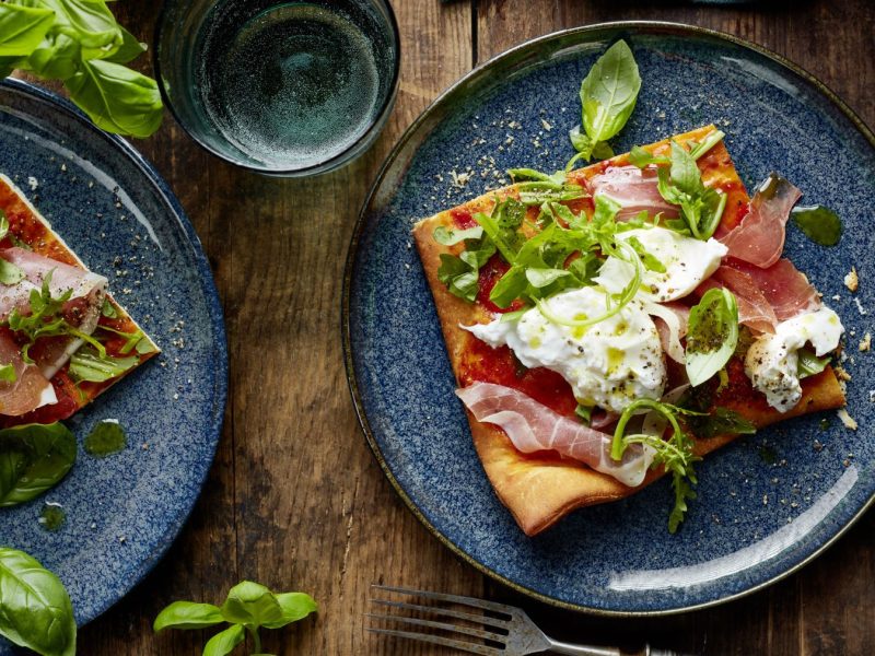 Pizza Crudo auf zwei grauen Tellern verteilt. Der Hintergrund ist hölzern, darauf Basilikum und ein Glas Wasser.