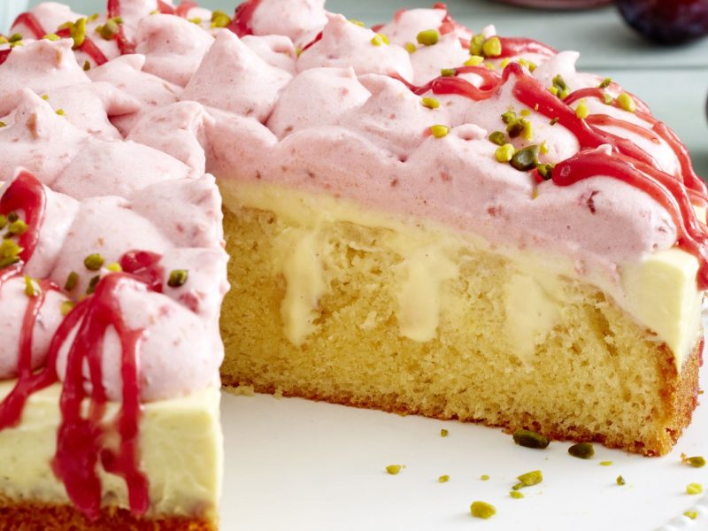 Draufsicht, schräg: Ein relativ flacher Kuchen, der auf einem hellen runden Untergrund liegt. Der Kuchen ist angeschnitten.