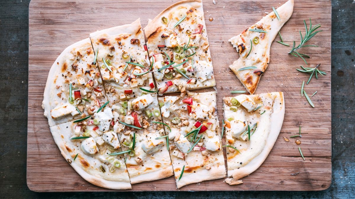 Knuspriger Flammkuchen mit Ziegenkäse, Apfel, Walnüssen und Frühlingszwiebeln, garniert mit Rosmarin auf einem Holzbrett.