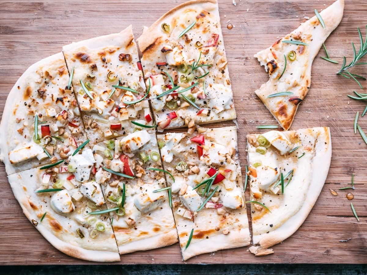 Knuspriger Flammkuchen mit Ziegenkäse, Apfel, Walnüssen und Frühlingszwiebeln, garniert mit Rosmarin auf einem Holzbrett.
