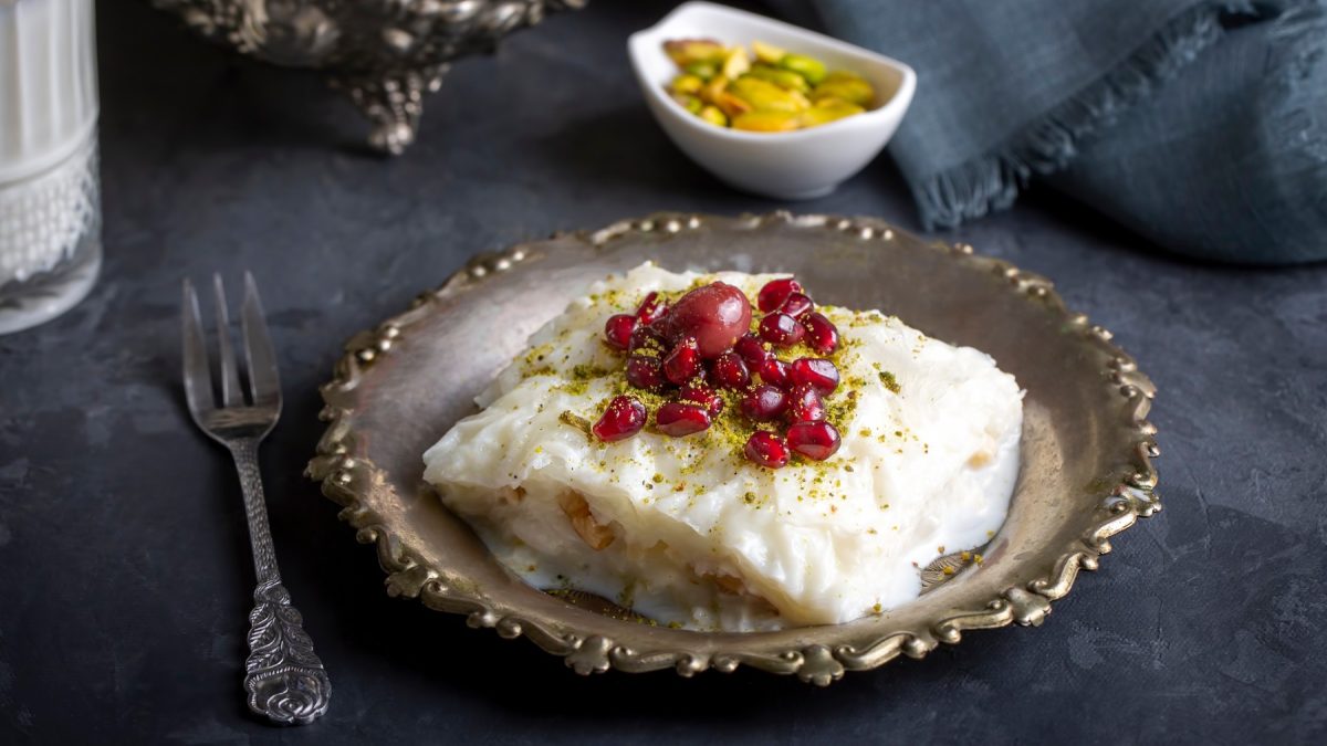 Türkisches Güllaç mit Granatapfel und Pistazien auf verziertem Metallteller, dunkler Untergrund, silberne Gabel.