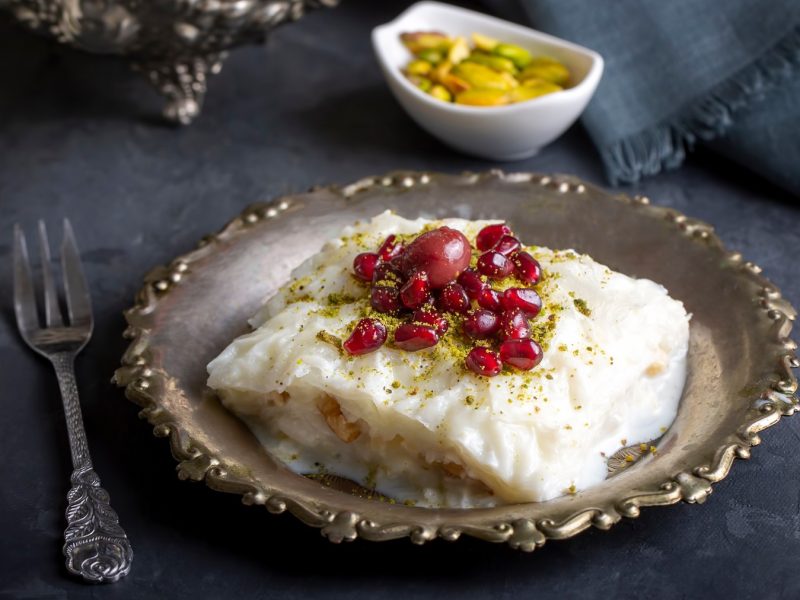 Türkisches Güllaç mit Granatapfel und Pistazien auf verziertem Metallteller, dunkler Untergrund, silberne Gabel.