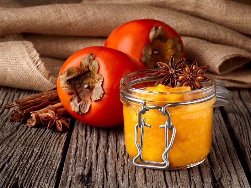 Eimn Glas mit der MArmelade auf einem dunklen Holztisch mit Kaki und Zimtstangen im Hintergrund.