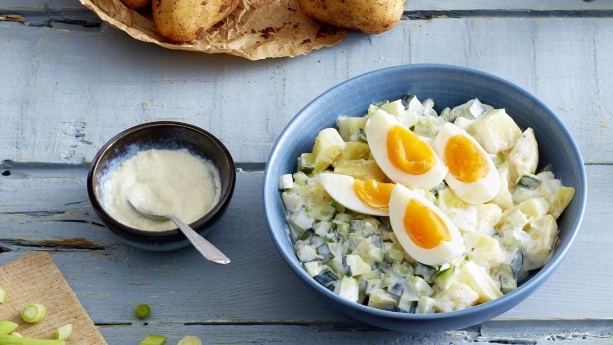 Cremiger Kartoffelsalat mit Gurken und Frühlingszwiebeln, garniert mit halbierten weichgekochten Eiern in blauer Schale.
