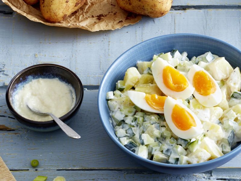 Cremiger Kartoffelsalat mit Gurken und Frühlingszwiebeln, garniert mit halbierten weichgekochten Eiern in blauer Schale.