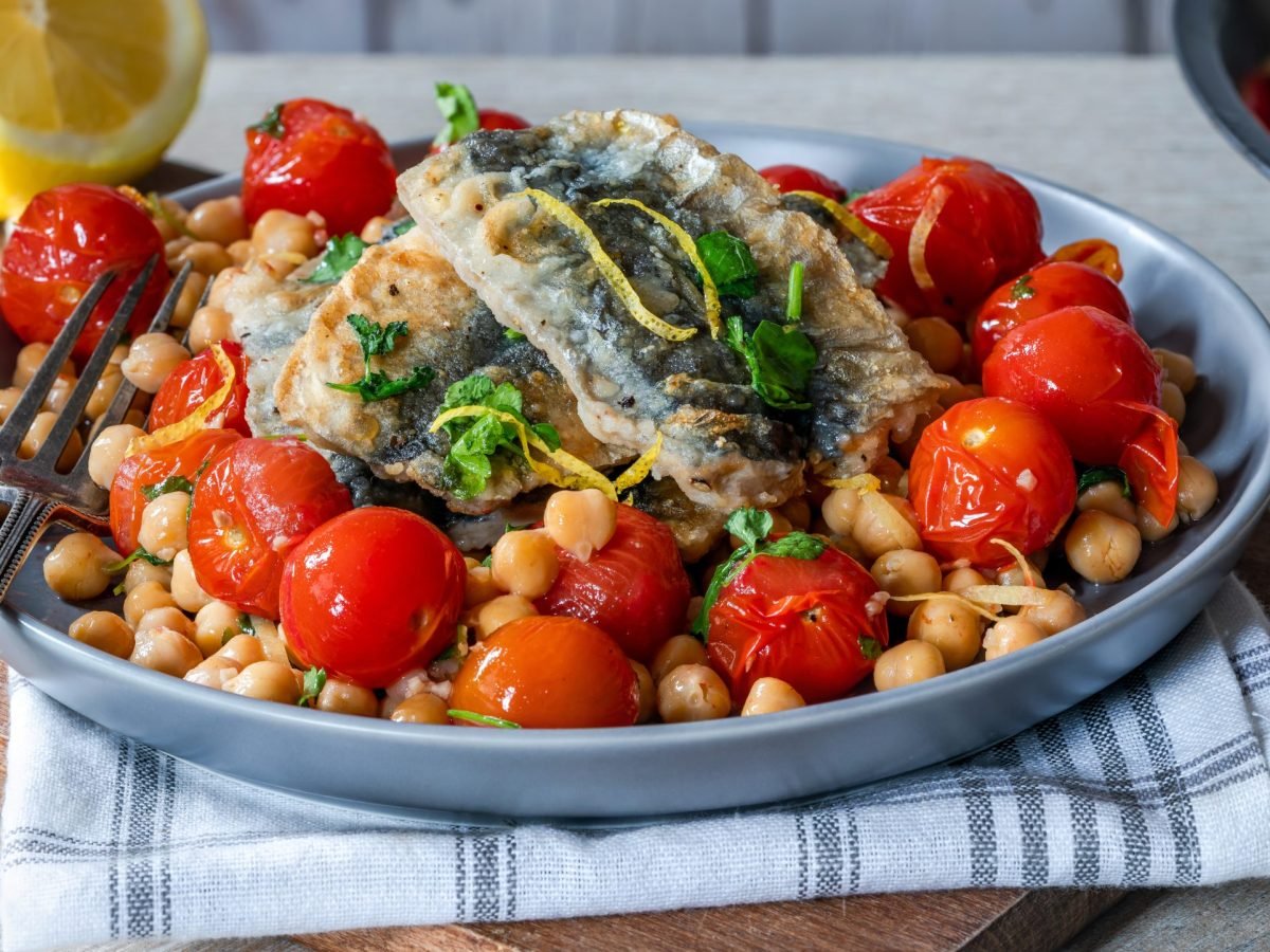 Knusprig gebratener Fisch mit Zitronenzesten auf einem Bett aus Kichererbsen und gerösteten Tomaten in einer grauen Schale.