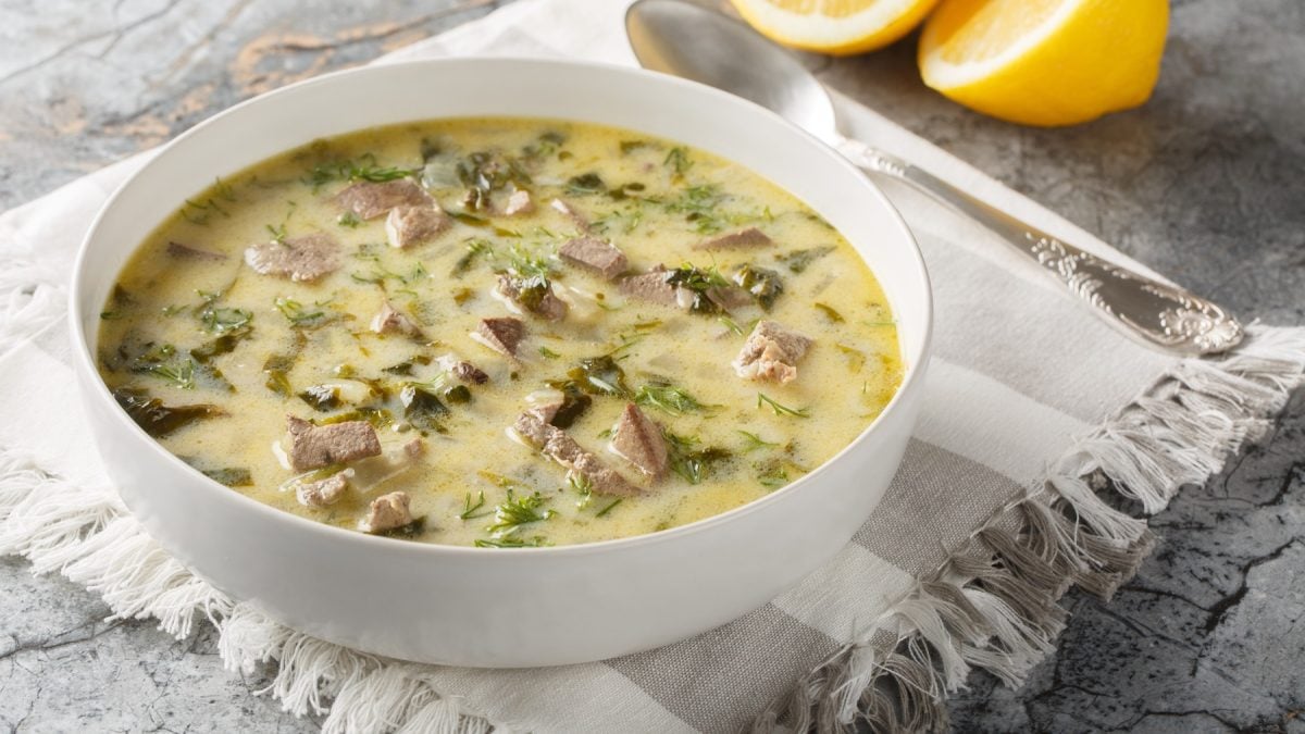 Eine Schale mit Magiritsa, zartem Fleisch und frischen Kräutern steht auf einem weißen Stofftuch, daneben zwei Zitronen.