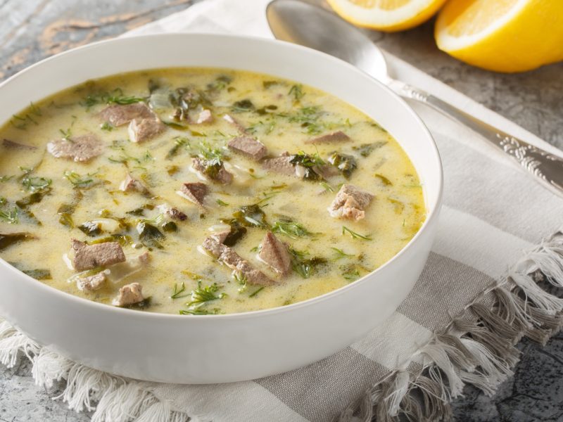 Eine Schale mit Magiritsa, zartem Fleisch und frischen Kräutern steht auf einem weißen Stofftuch, daneben zwei Zitronen.