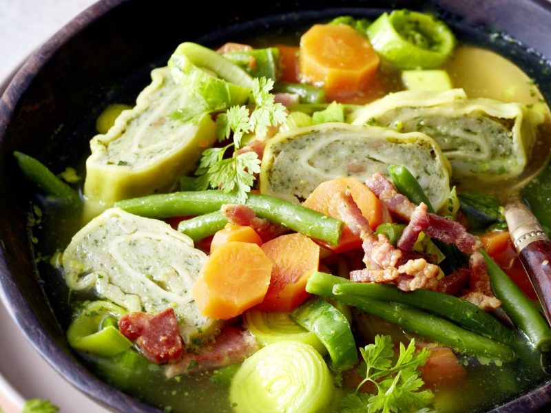 Bunte Suppe mit geschnittenen Maultaschen, Möhren, grünen Bohnen, Lauch und Speck, angerichtet in rustikaler Schale.