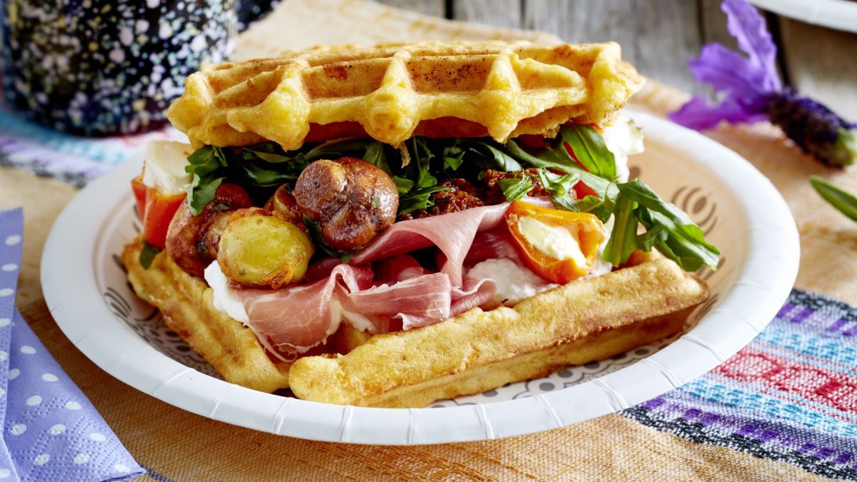 Herzhaft belegte Möhren-Waffeln mit Rucola, Schinken, gebratenen Pilzen, Tomatenhälften und cremigem Käse, serviert auf bunter Tischdecke.