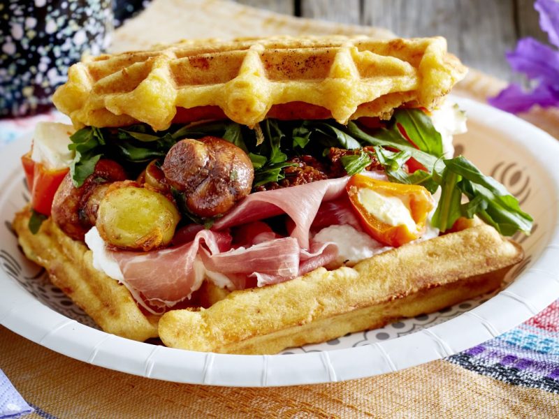 Herzhaft belegte Möhren-Waffeln mit Rucola, Schinken, gebratenen Pilzen, Tomatenhälften und cremigem Käse, serviert auf bunter Tischdecke.