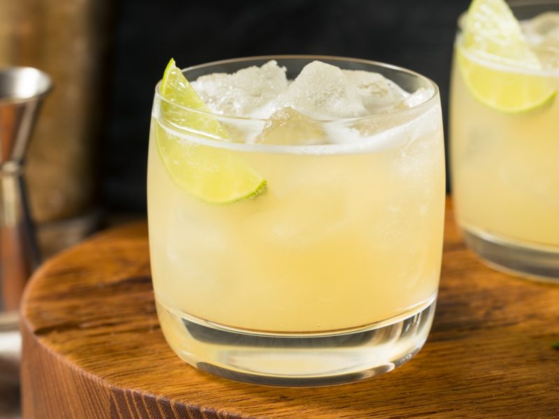 Zwei Gläser mit dem leicht gelben Cocktail auf einem Holzbrett mit Bar-Equipment im Hintergrund.