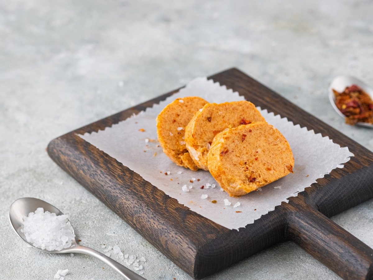 Drei Scheiben Paprika-Knoblauch-Butter mit grobem Meersalz auf dunklem Holzbrett mit Backpapier, daneben zwei silberne Löffel.