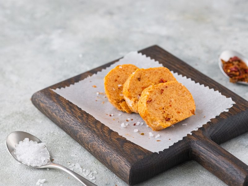 Drei Scheiben Paprika-Knoblauch-Butter mit grobem Meersalz auf dunklem Holzbrett mit Backpapier, daneben zwei silberne Löffel.