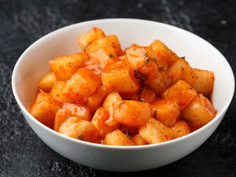 Ein weißes Schälchen mit dem Rettich Kimchi auf einem dunklen Untergrund. Alles nah und von der Seite fotografiert.