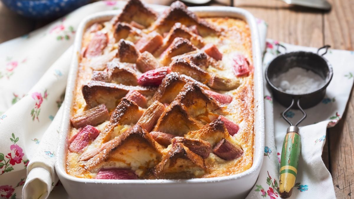 Süßer Rhabarber-Brotpudding mit karamellisierten Spitzen in weißer Form auf rustikalem Holztisch mit besticktem Tuch.