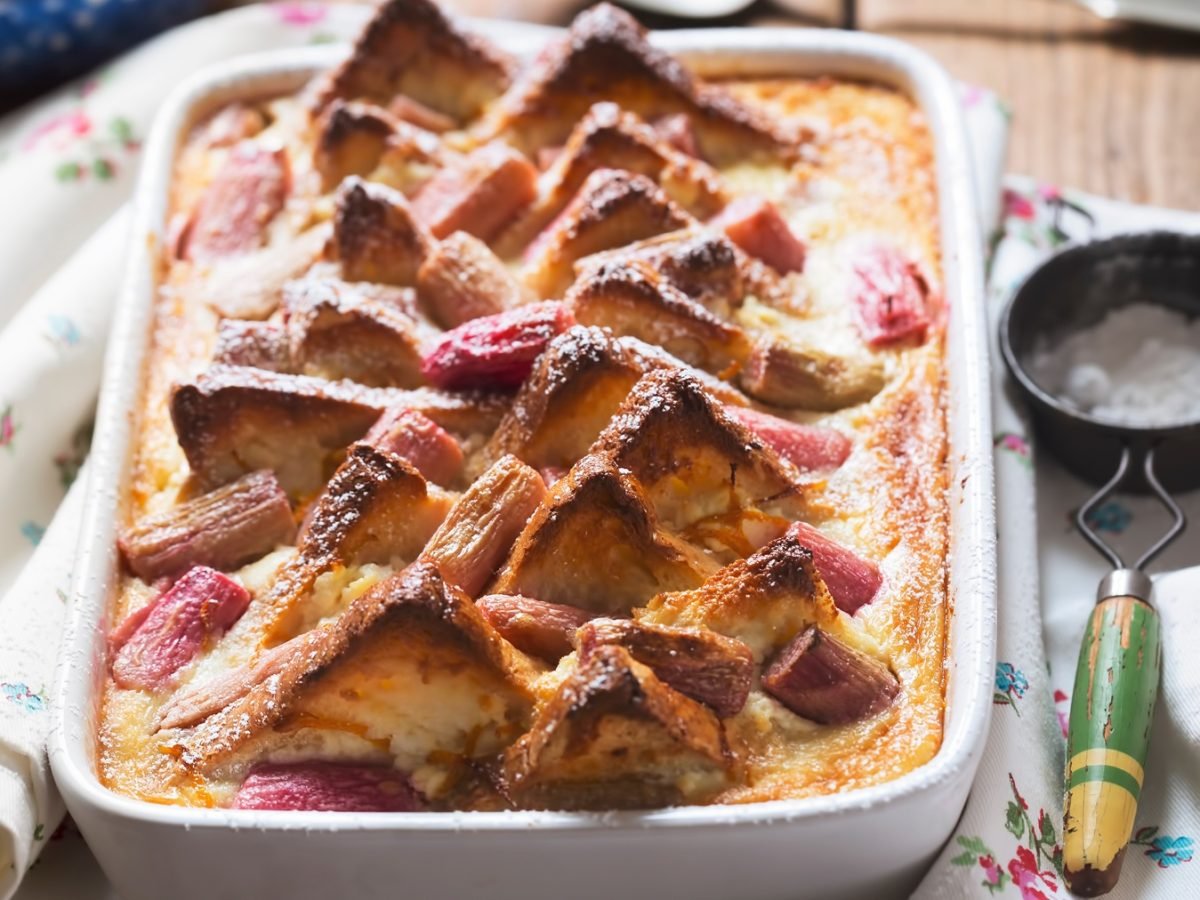Süßer Rhabarber-Brotpudding mit karamellisierten Spitzen in weißer Form auf rustikalem Holztisch mit besticktem Tuch.