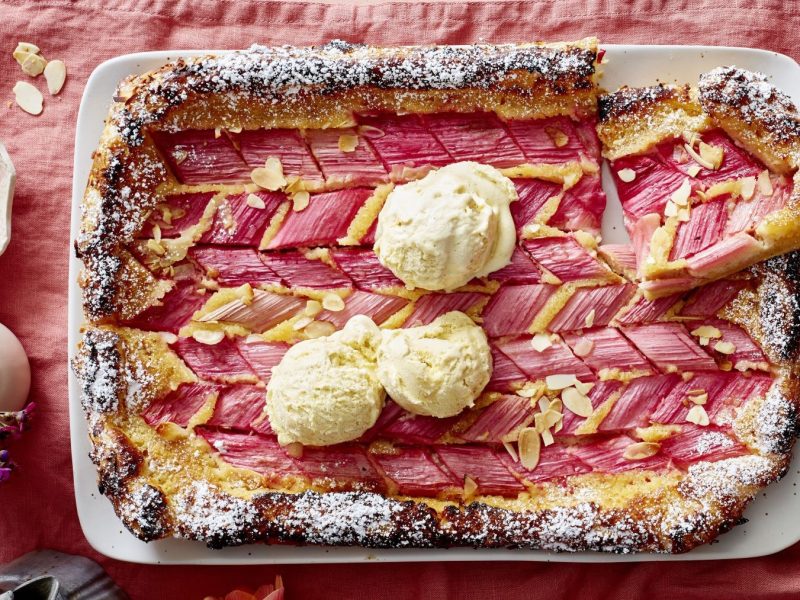 Ein rechteckiger weißer Teller mit der Rhabarber-Mandel-Galette auf einem Tisch mit rosafarbener Tischdecke. Alles direkt von oben fotografiert.