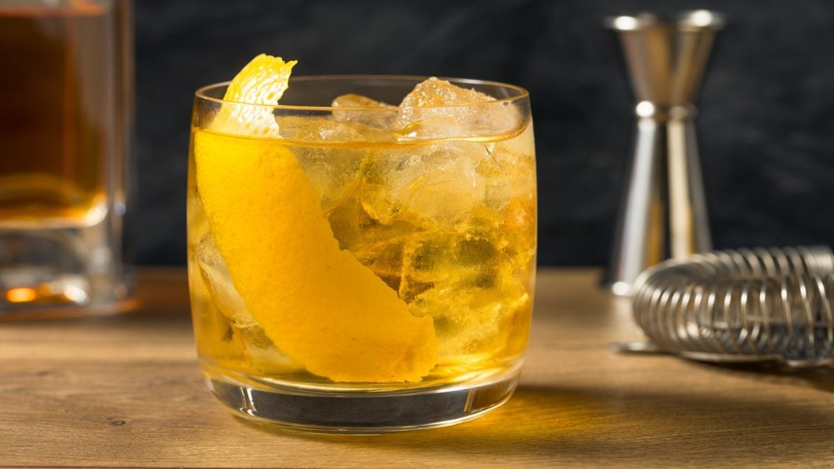 Bernsteinfarbener Rusty Nail mit Eis und Zitronenschale in einem Tumbler, daneben Barmesser und Messbecher aus Metall.