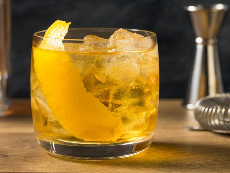 Bernsteinfarbener Rusty Nail mit Eis und Zitronenschale in einem Tumbler, daneben Barmesser und Messbecher aus Metall.
