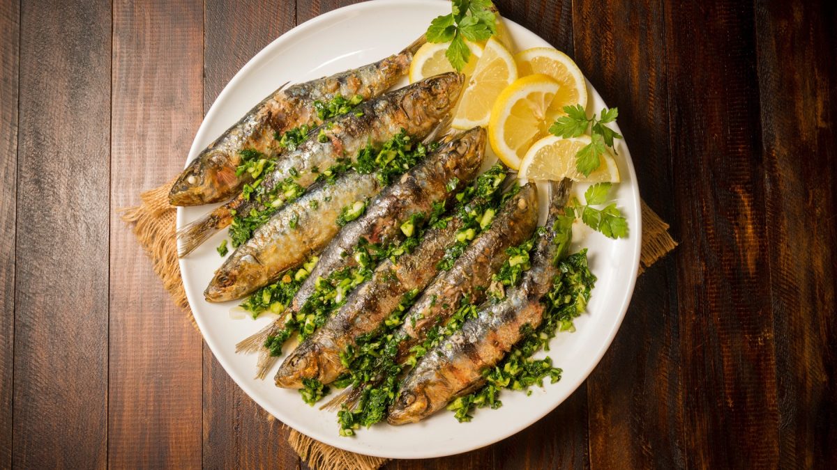 Sardinas a la plancha mit Kräutermarinade und Zitronenscheiben auf einem weißen Teller, serviert auf dunklem Holztisch.