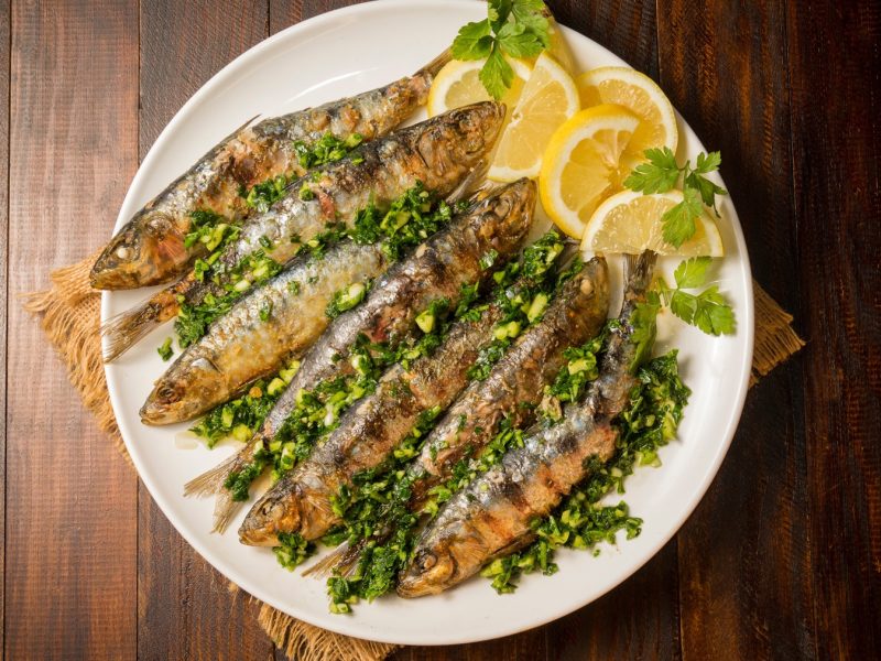Sardinas a la plancha mit Kräutermarinade und Zitronenscheiben auf einem weißen Teller, serviert auf dunklem Holztisch.