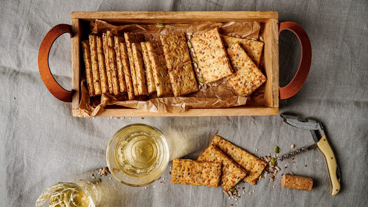 Rustikale Sauerteig-Discard-Cracker mit Körnermix und goldener Kruste neben Weißwein und geöffnetem Korken auf grauem Leinen.